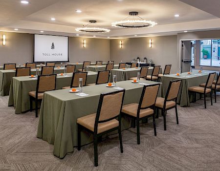 A conference room set up for a meeting, with long green tablecloths, rows of chairs, a projector screen at the front, and ambient ceiling lights.