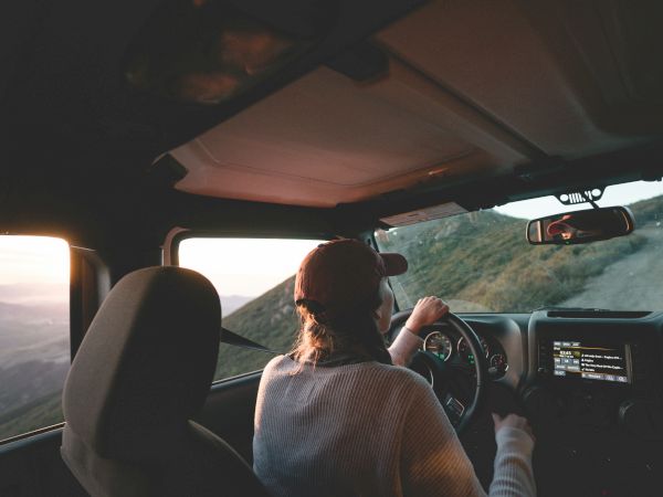 A person driving a car on a mountain road at sunset, seen from the back seat, with rugged scenery streaming by through the windows.