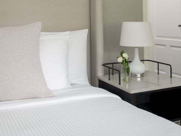 A tidy hotel bedroom with a white bed, a bedside table, a white lamp, and a small vase of flowers on the table, creating a calm, modern vibe.