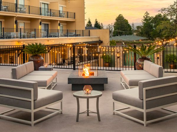 A stylish hotel patio with modern gray seating, a fire pit centerpiece, warm lighting, and potted palms at sunset, inviting and cozy.