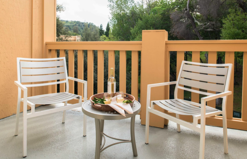 Two white patio chairs with a small round table on a balcony, drinks and food set out, railing and greenery in the background, relaxing scene.