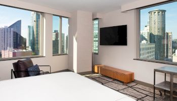 A modern hotel room with a large bed, wall-mounted TV, armchair, and cityscape views through large windows, featuring tall buildings outside.