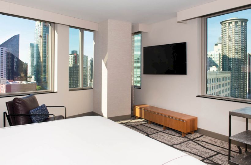 A modern hotel room with a large bed, wall-mounted TV, armchair, and cityscape views through large windows, featuring tall buildings outside.