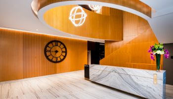 A modern reception area with a marble desk, large wall clock, spiral staircase, and hanging spherical lights, featuring wooden wall panels and floor tiles.