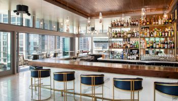 A modern bar area with a stylish wooden counter, high chairs, shelves stocked with various spirits, and large windows offering a city view.