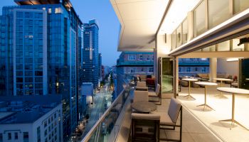 A modern cityscape viewed from a high balcony at dusk, featuring tall buildings, streetlights, and a well-furnished outdoor seating area.