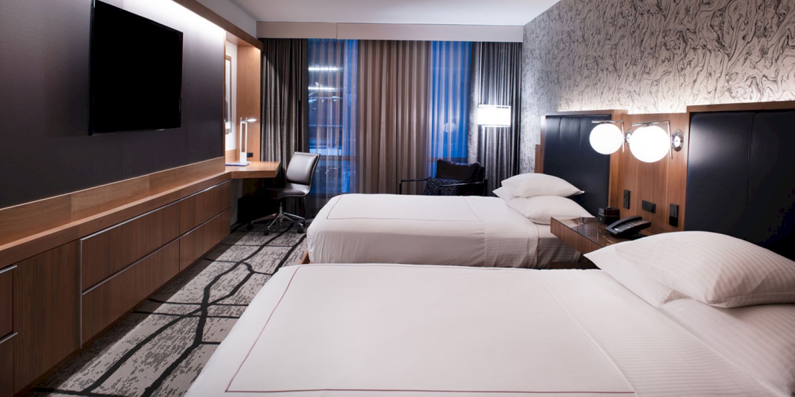 The image shows a modern hotel room with two neatly made beds, a flat-screen TV, a desk with a chair, and contemporary decor.