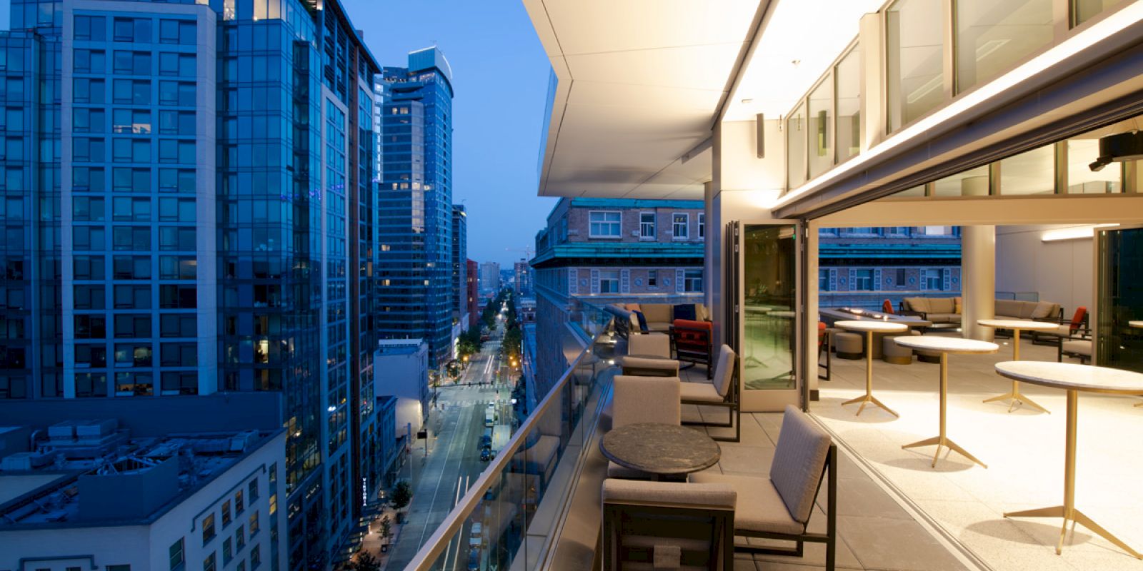 A modern rooftop terrace with outdoor seating, tables, and cityscape views at dusk. High-rise buildings are visible in the background.
