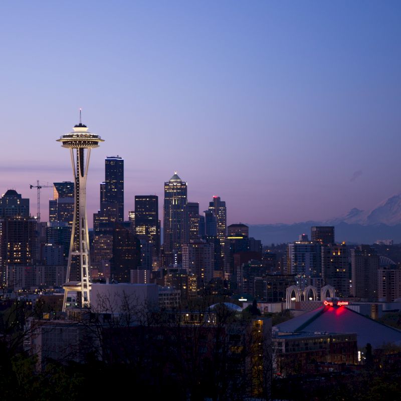 A cityscape image of Seattle, featuring the Space Needle and surrounding skyscrapers at dusk, with Mount Rainier visible in the background.