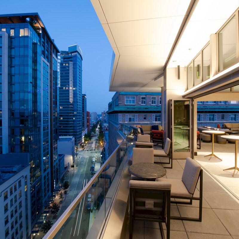 The image shows a modern cityscape view from a balcony with outdoor seating and tables, overlooking buildings and a street at dusk.