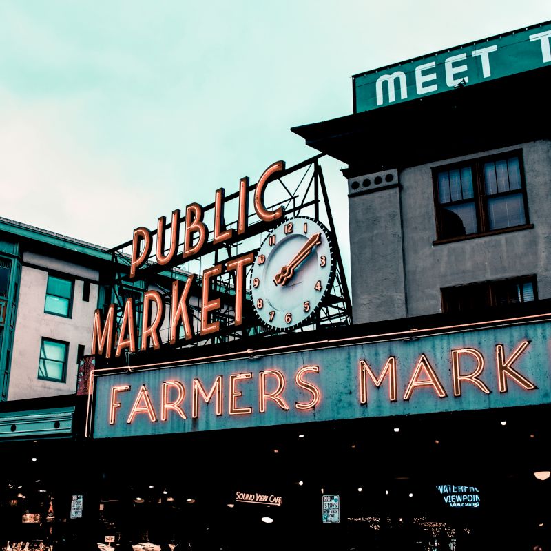 This image shows a neon sign for a public farmers market with a clock above, situated among older style buildings.