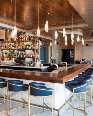 This image shows a modern bar with a well-stocked liquor display, hanging lights, blue bar stools, and a seating area to the right.