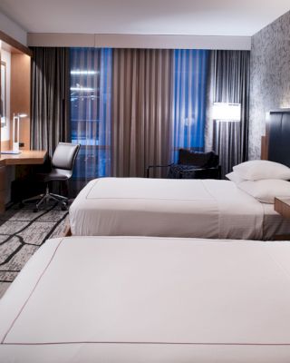 This image shows a modern hotel room with two beds, a desk, a flat-screen TV, and contemporary decor, featuring wood and patterned wallpaper.