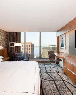 A modern hotel room with a large bed, a desk with a chair, a wall-mounted TV, and a view of the city through floor-to-ceiling windows.