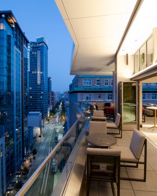 A cityscape view at dusk from a modern outdoor terrace, featuring seating areas and buildings with tall glass windows lining a well-lit street.