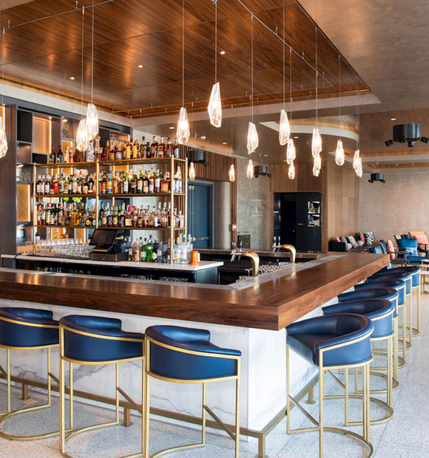 A modern bar with a wooden counter, blue bar stools, hanging lights, and a well-stocked liquor shelf. The interior is spacious and stylish.