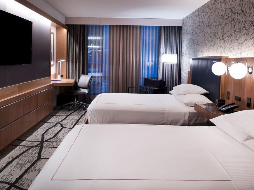 This image shows a modern hotel room with two beds, a TV, a work desk, and large windows with curtains, featuring stylish decor and lighting.