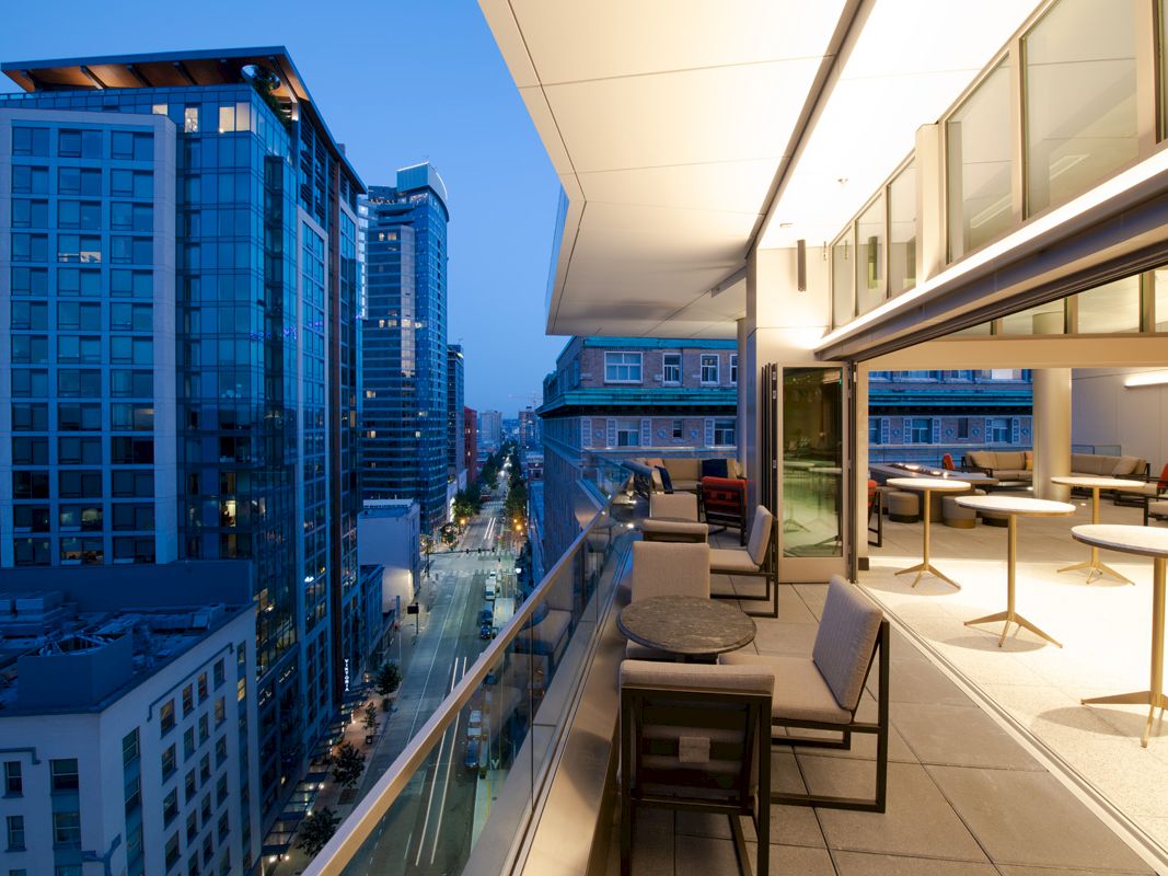 A modern rooftop terrace at dusk with tables, chairs, and a glass railing, overlooking a city street lined with tall buildings.