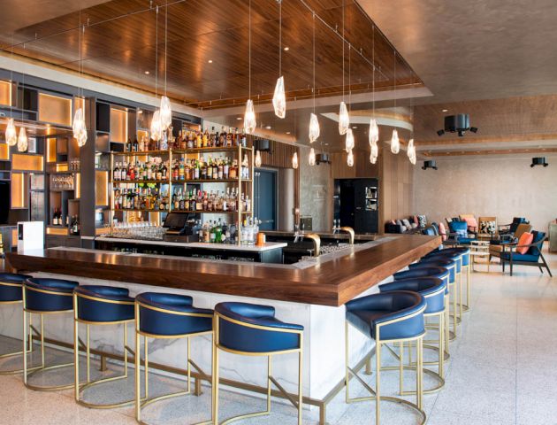 A modern bar with wooden countertops, shelves stocked with various bottles, blue bar stools, hanging lights, and a spacious seating area.