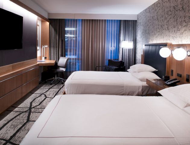 The image shows a modern hotel room with two neatly made beds, a large TV mounted on the wall, a desk with a chair, and stylish decor.