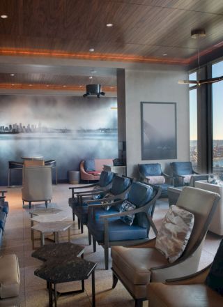 A modern lounge with seating, large windows with a waterfront view, and a cityscape mural on the wall.