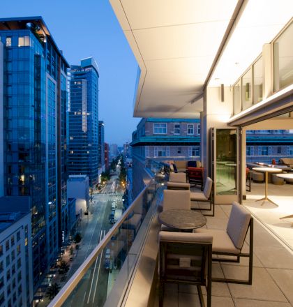 A modern urban balcony at dusk overlooks a street lined with tall buildings, featuring outdoor seating and a partially open indoor area.