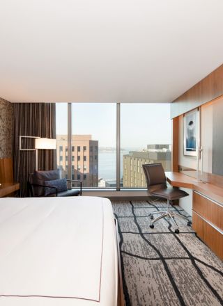 A modern hotel room with a large bed, a desk, a chair, a wall-mounted TV, and a view of buildings and water through a large window.