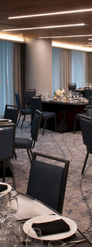 A banquet hall set up for an event with round tables, dark chairs, elegant place settings, and soft lighting from overhead fixtures.