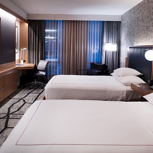 A modern hotel room with two double beds, a wall-mounted TV, a desk with a chair, bedside tables with lamps, and contemporary decor.