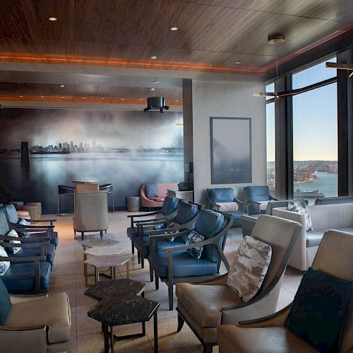 A modern lounge area with numerous chairs, a scenic wall mural, and large windows offering a view of a Ferris wheel and waterfront.