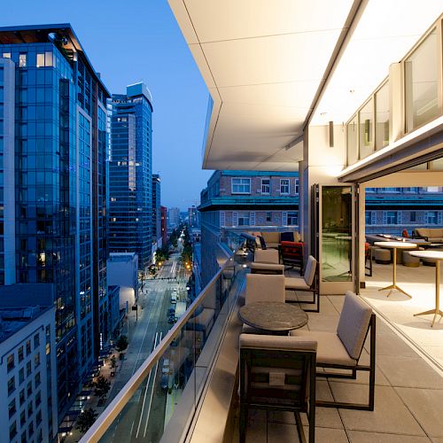 This image shows a modern urban balcony at dusk, overlooking a city street lined with high-rise buildings, featuring tables, chairs, and indoor seating.