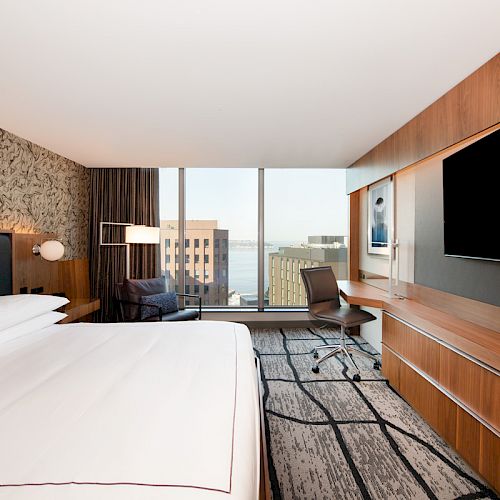 The image shows a modern hotel room with a large bed, a wall-mounted TV, a desk, and floor-to-ceiling windows offering a city view.