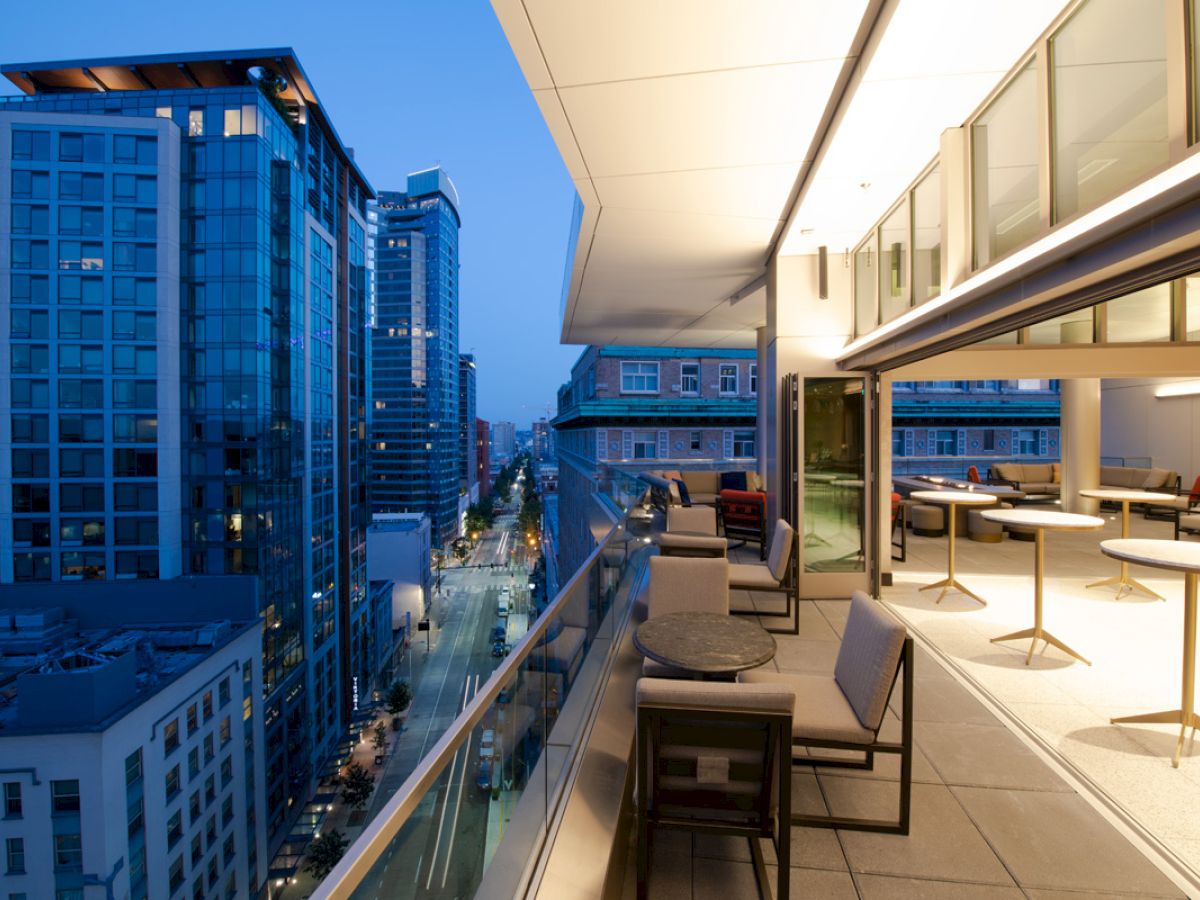 The image shows a modern balcony with seating arrangements overlooking a cityscape at dusk, with tall buildings and streets visible below.