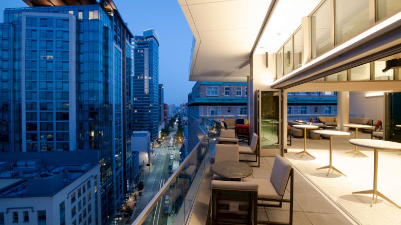 The image shows a modern balcony with seating arrangements overlooking a cityscape at dusk, with tall buildings and streets visible below.