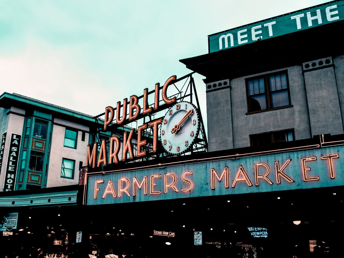 The image shows the iconic neon signs of the Public Market and Farmers Market at Pike Place Market in Seattle, Washington, with buildings in the background.