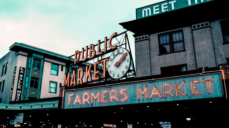 The image shows the iconic neon signs of the Public Market and Farmers Market at Pike Place Market in Seattle, Washington, with buildings in the background.