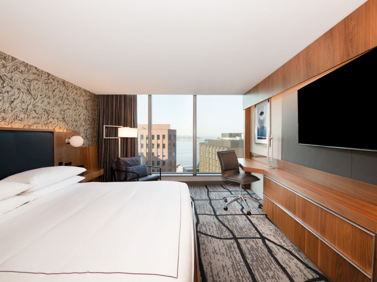 A modern hotel room with a large bed, desk, chair, and TV, features a window with a city view and contemporary decor.