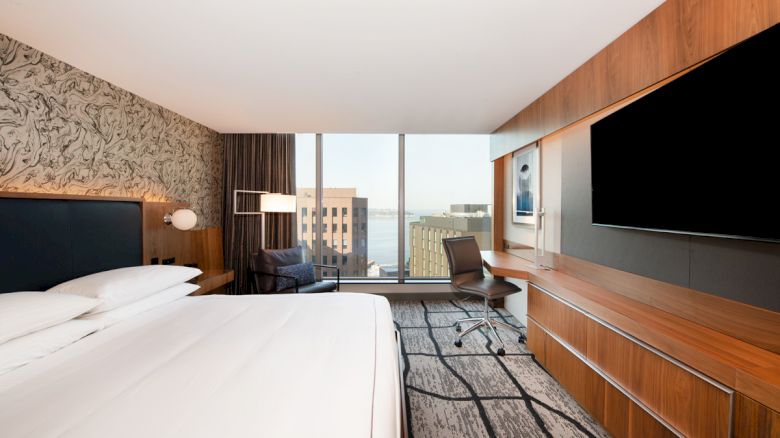 A modern hotel room with a large bed, desk, chair, and TV, features a window with a city view and contemporary decor.