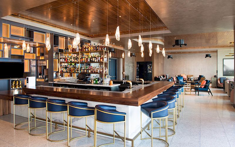 This image shows a modern bar with a variety of liquor bottles, blue bar stools, hanging pendant lights, and a lounge area in the background.