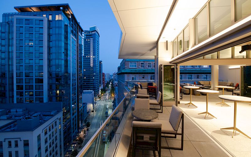 The image shows a modern balcony with seating overlooking a cityscape of tall buildings during the evening, with a view of a street below.