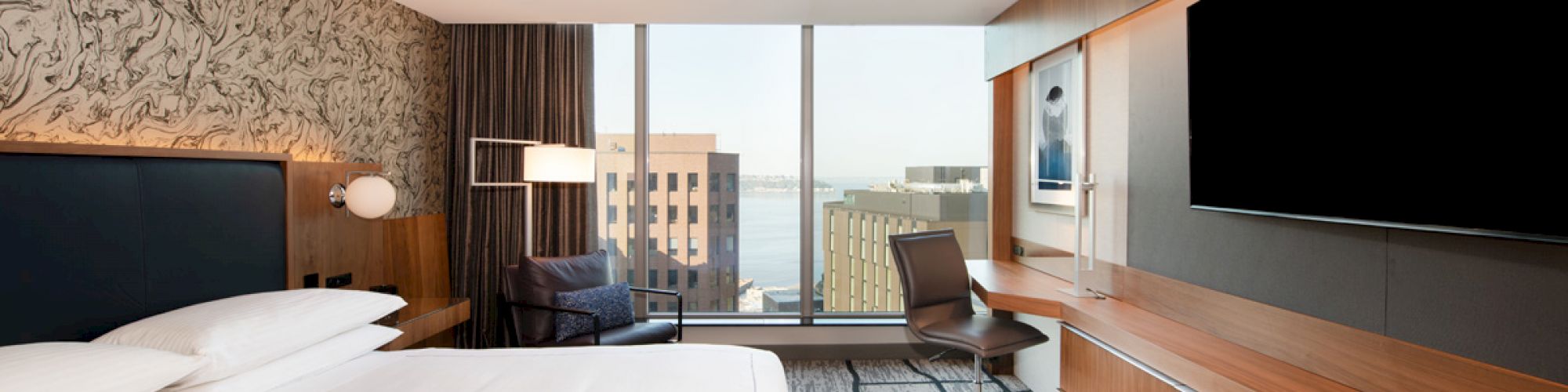 A modern hotel room features a large bed, a wall-mounted TV, a desk with a chair, and a view of city buildings through a large window.