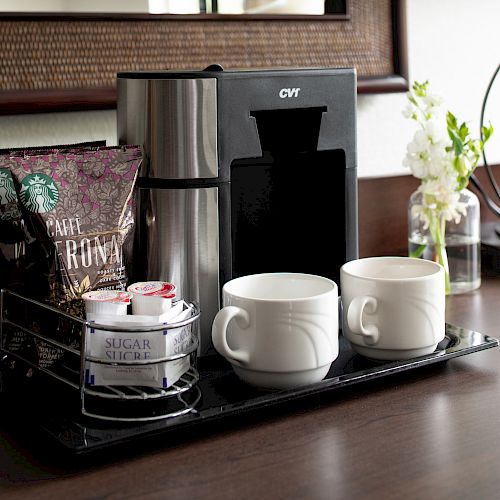 The image shows a coffee setup with a coffee maker, Starbucks coffee packets, sugar, cream, and two white mugs on a tray, next to a vase.