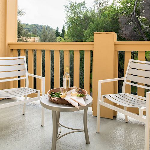 Two white patio chairs and a small table with a tray, bottle, and glasses are on a balcony with a forested background.