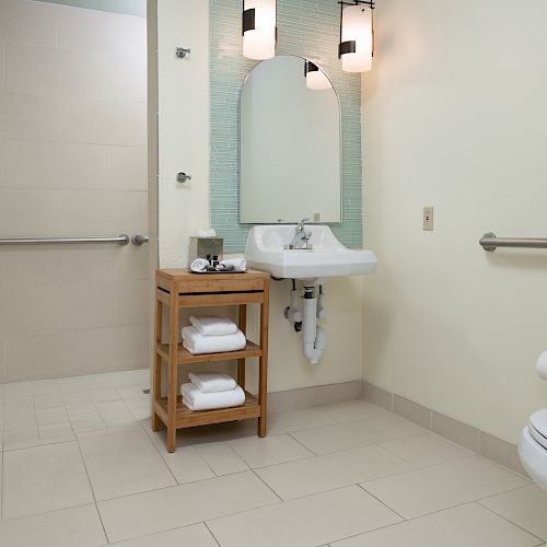 The image depicts a clean, modern bathroom with a sink, wooden shelving unit, toilet, and shower. Wall-mounted lamps and towel racks are present.