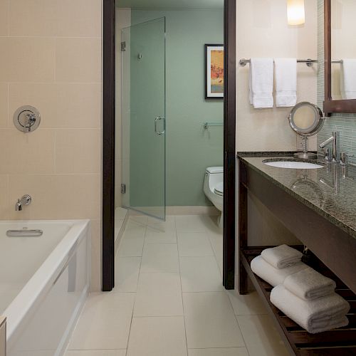 The image shows a clean, modern bathroom with a bathtub, double sinks, a large mirror, a glass-enclosed shower, and neatly arranged towels.