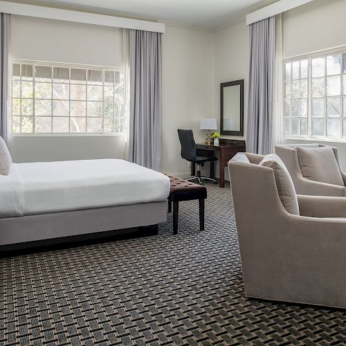 The image shows a well-lit hotel room with a bed, armchairs, a desk, and large windows. The room has neutral decor and a patterned carpet.