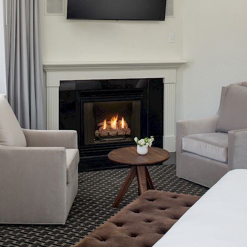 A cozy room features two beige chairs, a small wooden table, a fireplace, and a TV mounted above it.