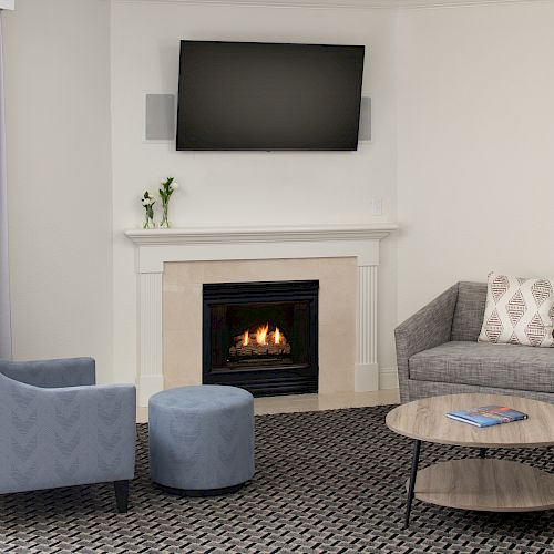 A cozy living room with a gray couch, a blue chair, a matching ottoman, a round coffee table, a fireplace, and a wall-mounted TV above it.