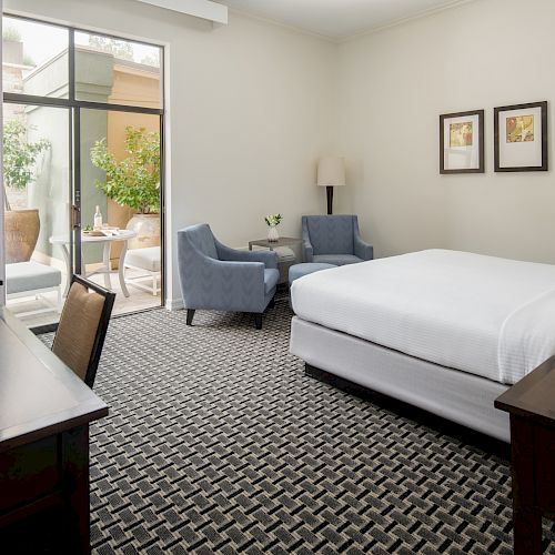 A neatly arranged hotel room with a bed, two chairs, a desk with a TV, and a sliding door leading to an outdoor patio area.