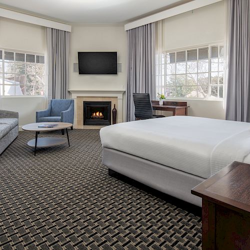 A spacious hotel room with a large bed, a sofa, armchair, TV, fireplace, and study desk, decorated in neutral tones with light gray curtains.